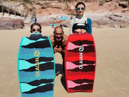 Drei Personen stehen am Strand, halten bunte Bodyboards und lächeln fröhlich in die Kamera, umgeben von einer schönen Landschaft.