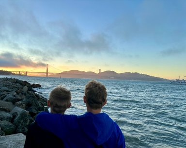 Zwei Jungen stehen am Wasser und blicken auf die Golden Gate Bridge, während die Sonne hinter den Hügeln untergeht.