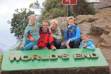 Eine Familie posiert fröhlich vor dem Schild 'WORLD'S END' in der Natur, umgeben von grünen Pflanzen und Felsen.