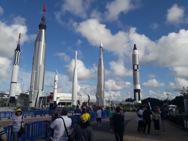 Eine Gruppe von Besuchern steht vor mehreren beeindruckenden Raketen im Kennedy Space Center unter einem blauen Himmel.