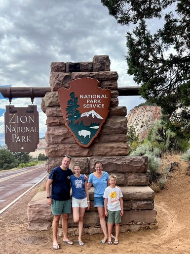 Eine Familie steht lächelnd vor dem Eingang zum Zion Nationalpark, umgeben von beeindruckenden Felsen und Bäumen.