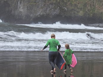 Eine Frau und ein Mädchen in grünen Neoprenanzügen gehen mit Bodyboards am Strand entlang, während die Wellen brechen.