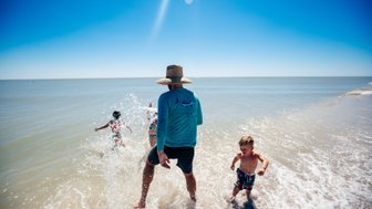 Ein Erwachsener mit einem Hut steht im Wasser, während zwei Kinder fröhlich im Hintergrund spielen und spritzen.