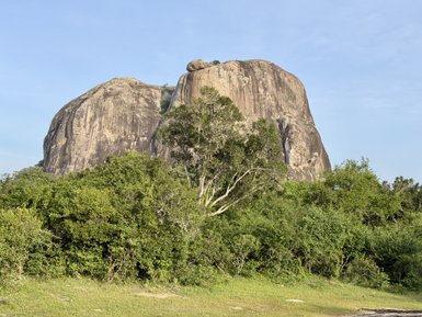 Ein majestätischer Felsen erhebt sich über üppiges, grünes Gestrüpp unter einem klaren blauen Himmel.