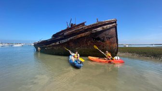 Zwei Kajakfahrer paddeln in ruhigem Wasser neben einem alten, verrosteten Schiff, das am Ufer liegt.
