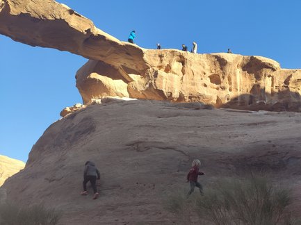 Zwei Kinder klettern an einer steilen Felswand in der Wüste Wadi Rum, während andere Menschen oben auf einem Felsen stehen.