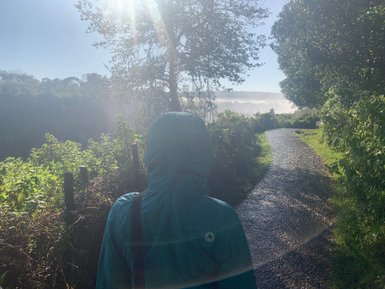 Eine Person in einer blauen Regenjacke steht auf einem schmalen Weg, umgeben von üppigem Grün und Sonnenlicht.