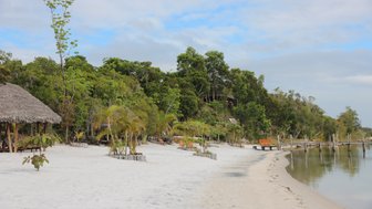 Ein ruhiger Strand mit feinem, weißem Sand, umgeben von üppigem Grün und einer Holzterrasse am Wasser.