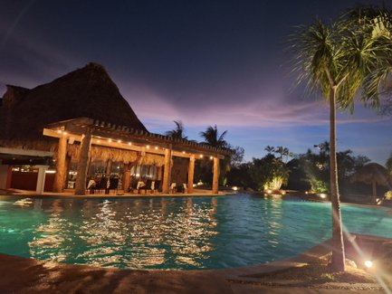 Ein einladender Poolbereich mit sanftem Wasser, umgeben von Palmen und einem strohgedeckten Pavillon bei Dämmerungslicht.