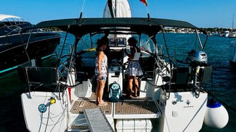 Zwei Frauen stehen auf einem Segelboot, das an einem Steg im Hafen festgemacht ist, umgeben von anderen Yachten.