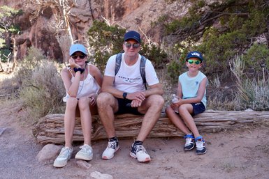 Eine Familie sitzt auf einem Baumstamm im Freien, umgeben von trockener Vegetation und Felsen, während sie sich ausruhen.