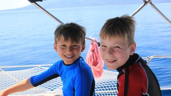 Zwei fröhliche Jungen in Neoprenanzügen sitzen auf einem Boot und genießen die Sonne und das Wasser um sie herum.