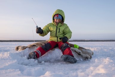 Ein Kind in einer grünen Jacke sitzt auf einem Schneehaufen und angelt durch ein Loch im Eis, umgeben von winterlicher Landschaft.