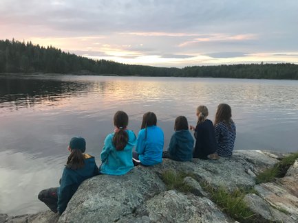Sechs Kinder sitzen auf einem Felsen am Ufer eines ruhigen Sees und beobachten den Sonnenuntergang über den Bäumen.