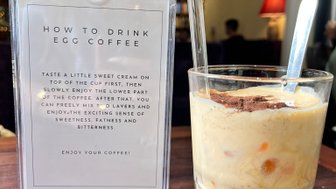 Ein Glas Eierkaffee mit einer Schicht süßer Sahne und einer dunklen Kaffeeschicht, daneben eine Anleitung zum Trinken.