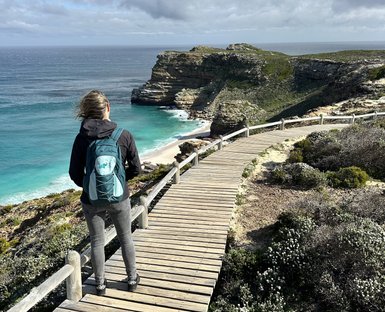 Eine Frau mit einem Rucksack steht auf einem Holzsteg und blickt auf die Küste mit türkisfarbenem Wasser und Klippen.