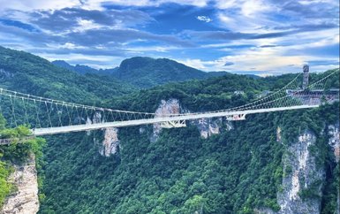 Eine beeindruckende Hängebrücke schwebt über eine grüne Schlucht, umgeben von majestätischen Bergen und einem strahlend blauen Himmel.