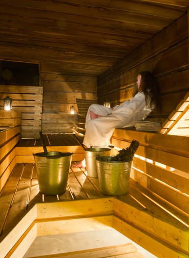 In einer warmen Sauna sitzt eine Person in einem weißen Bademantel entspannt auf einer Holzbank, umgeben von Eimern.