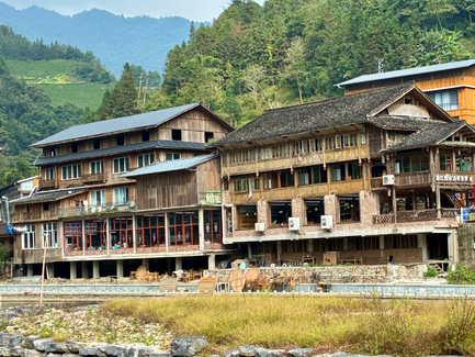 Holzhäuser mit traditioneller Architektur stehen am Ufer eines ruhigen Flusses, umgeben von grünen Bergen und sanften Hügeln.