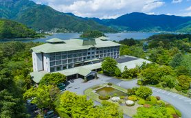 Ein modernes Hotel mit grünem Dach, umgeben von üppiger Natur und Blick auf einen ruhigen See und die Berge im Hintergrund.