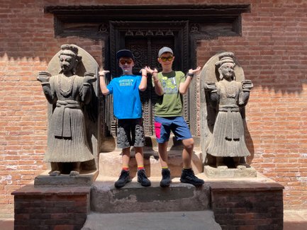 Zwei Jungen posieren fröhlich zwischen zwei beeindruckenden Steinfiguren in Bhaktapur, umgeben von rotem Ziegelmauerwerk.