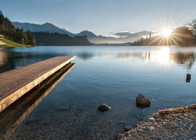 Ein ruhiger See spiegelt die umliegenden Berge und den Sonnenaufgang wider, während ein Holzsteg ins Wasser führt.