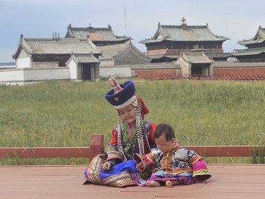 Zwei Kinder in traditioneller mongolischer Kleidung sitzen auf einem Holzdeck vor dem Erdene Zuu Kloster in der Natur.