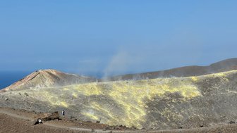 Ein beeindruckender Vulkan mit gelben Schwefelfeldern und rauchenden Ausläufern, umgeben von einer kargen Landschaft.