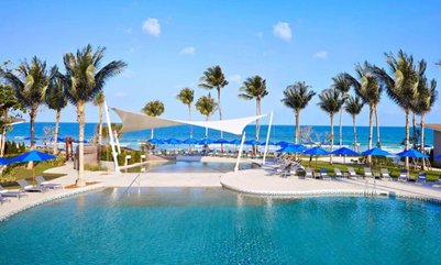 Ein glitzernder Pool umgeben von Palmen und blauen Sonnenschirmen, mit dem Meer im Hintergrund und strahlend blauem Himmel.