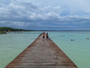 Ein Holzsteg führt über das türkisfarbene Wasser, während Menschen am Ufer und im Wasser entspannen.