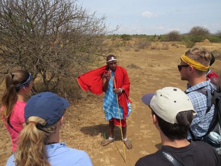Eine Gruppe von Touristen hört einem Maasai-Guide zu, der in traditioneller Kleidung auf einem trockenen Feld spricht.