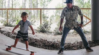 Ein kleiner Junge steht auf einem Skateboard und lächelt, während ein Erwachsener ihn ermutigend beobachtet.
