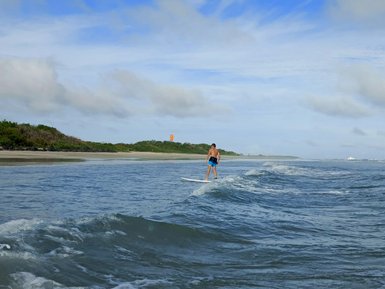 Ein junger Surfer steht auf seinem Brett und reitet sanfte Wellen an einem ruhigen Strand unter einem blauen Himmel.