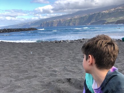 Ein Junge sitzt am Strand und gräbt mit seinen Händen im schwarzen Sand, während er auf das Meer blickt.