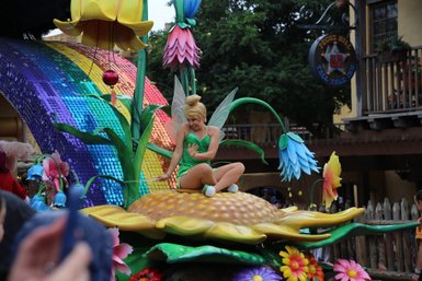Eine fröhliche Fee sitzt auf einer großen Blume, umgeben von bunten Blumen und glitzernden Regenbogenfarben.