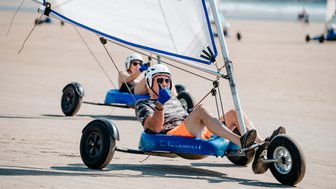 Ein Mann in einem blauen Strandbuggy mit einem Segel fährt über den Sand, während er Daumen hoch zeigt.