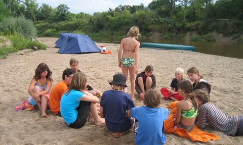 Eine Gruppe von Kindern sitzt im Sand am Flussufer, während sie an einer Campbesprechung teilnehmen und sich unterhalten.