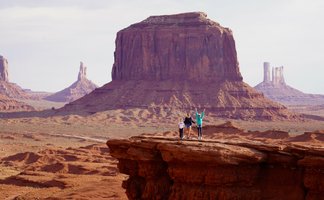 Drei Personen stehen auf einer Felsplattform in Monument Valley, umgeben von beeindruckenden roten Felsen und Hügeln.