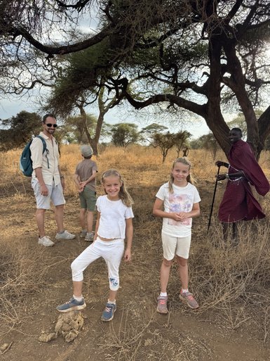 Eine Gruppe von Kindern und einem Erwachsenen posiert fröhlich in der trockenen Savanne, umgeben von hohen Bäumen.