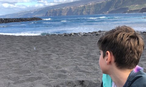 Ein Junge sitzt am Strand und gräbt mit seinen Händen im schwarzen Sand, während er auf das Meer blickt.