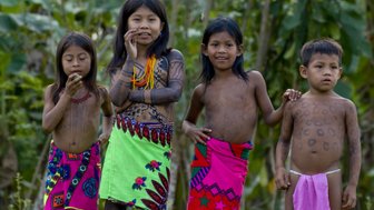 Vier Kinder in traditioneller Kleidung, die mit kulturellen Tätowierungen geschmückt sind, stehen lächelnd in der Natur.