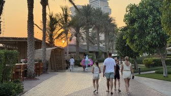 Eine Familie mit zwei Kindern spaziert auf einem gepflasterten Weg, umgeben von Palmen und einem Sonnenuntergang im Hintergrund.