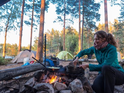 Eine Frau kocht am Lagerfeuer in einem Waldcamp, umgeben von Zelten und Bäumen, während die Sonne untergeht.
