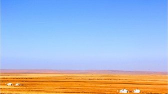 Eine weite, goldene Steppe erstreckt sich unter einem klaren blauen Himmel, mit wenigen Jurten in der Ferne.