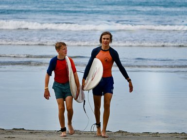 Zwei Surfer in Badesportkleidung gehen am Strand entlang, tragen ihre Surfbretter und haben grinsende Gesichter.