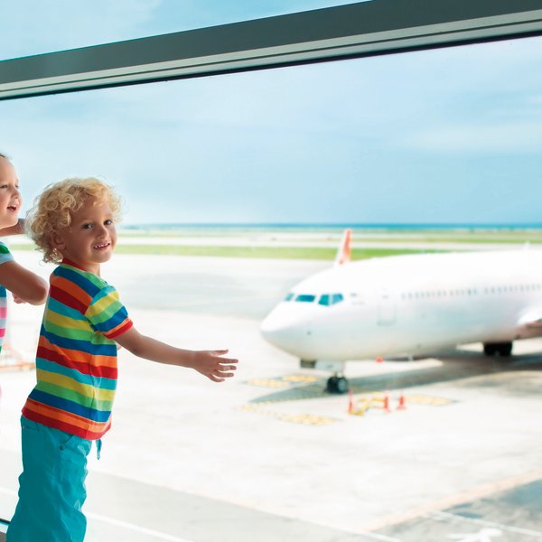 Zwei fröhliche Kinder stehen am Fenster eines Flughafens und beobachten ein Flugzeug auf dem Rollfeld.