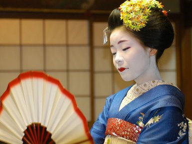 Eine Geisha in traditioneller Kleidung hält einen Fächer und zeigt eine nachdenkliche Miene in einem japanischen Raum.