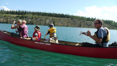 Eine Familie paddelt in einem roten Kanu auf einem klaren, blauen Gewässer, umgeben von grünen Bäumen und Hügeln.