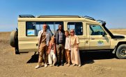 Eine Familie steht lächelnd vor einem Geländewagen in der afrikanischen Savanne, umgeben von einer weiten, trockenen Landschaft.