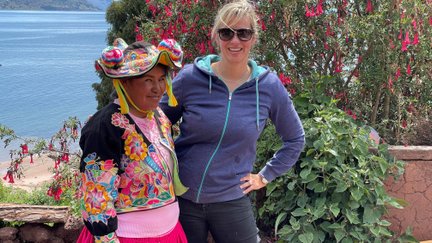 Eine einheimische Frau in traditioneller Kleidung steht neben einer Frau mit Sonnenbrille, beide lächeln vor einer malerischen Kulisse.